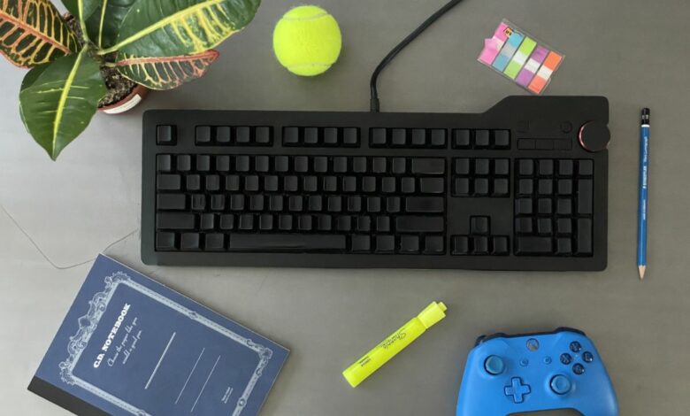 A black, unlabeled keyboard surrounded by other common objects including an Xbox controller and tennis ball.